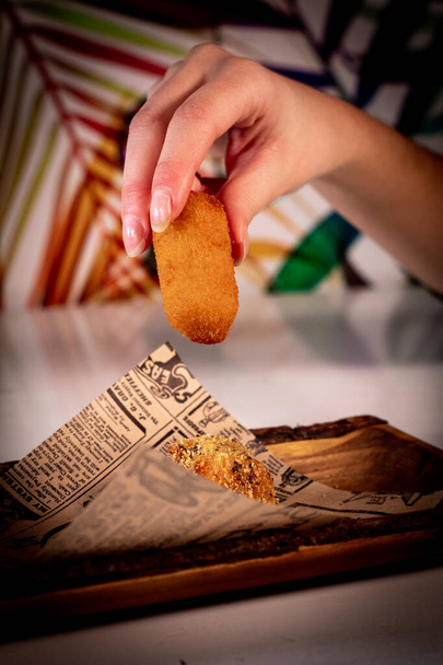 Eine feminine Hand, die ein traditionelles spanisches Kroketten-Essen probiert, das auf einem Tablett mit Briefpapier präsentiert wird. Bunte Pflanzen im Boden. - Foto, Bild