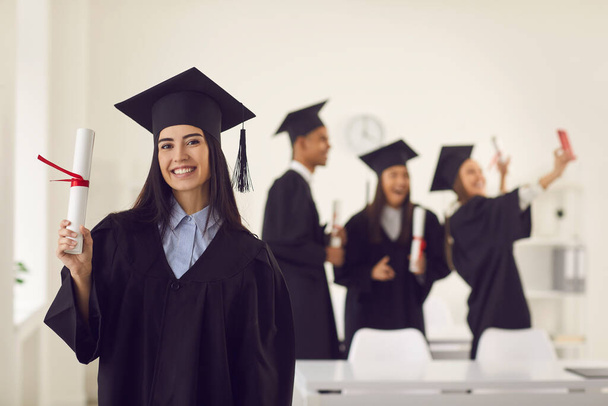 Female graduate shows the camera a diploma in her hands while standing in the classroom. - Φωτογραφία, εικόνα