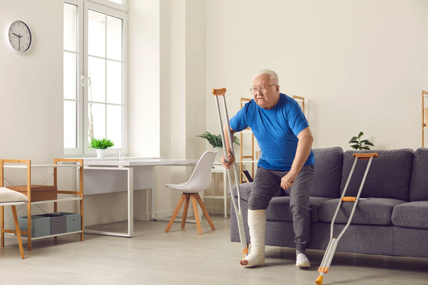 Senior man with broken leg trying to stand up from sofa and walk with crutches at home - Valokuva, kuva