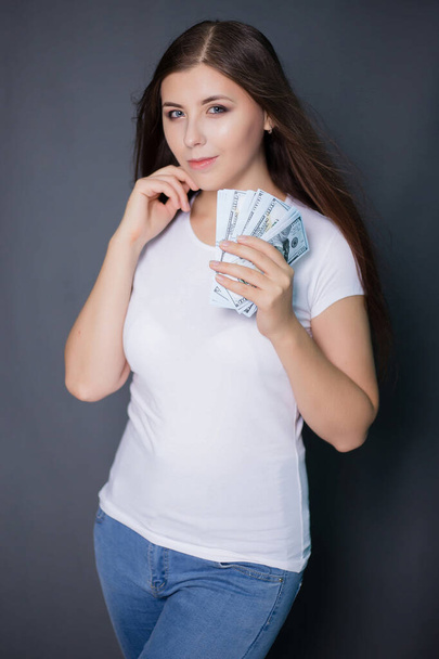 young, beautiful woman in a white T-shirt is holding dollars in hands. Studio photo on a gray background. - Foto, Imagem