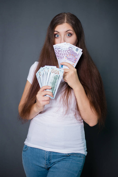 young, beautiful woman in white t-shirt and blue jeans holds euro and dollars in hands. Studio photo on a gray background. - Foto, afbeelding