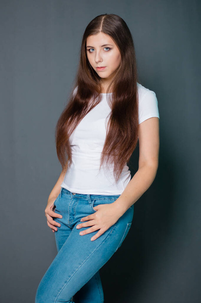 Young, beautiful woman in a white T-shirt and blue jeans. Studio photo on a gray background. - Фото, зображення