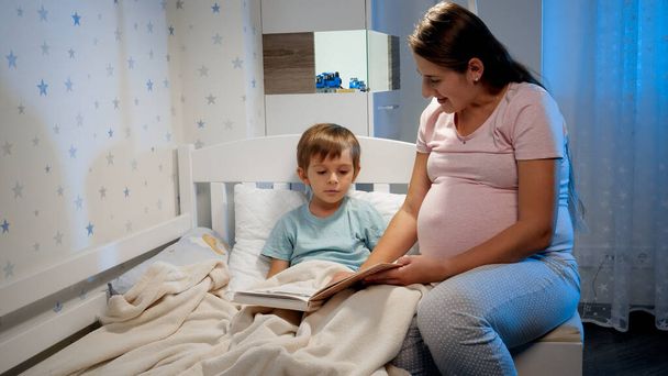 Hermosa madre embarazada readng libro de cuentos a su hijo mayor acostado en la cama por la noche antes de ir a dormir. Padres leyendo a los niños - Foto, imagen