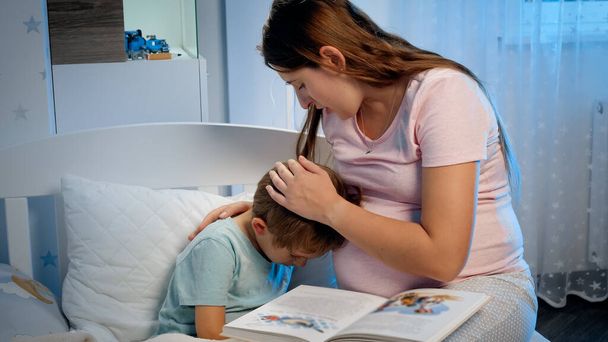 Молодая беременная мать обнимает и успокаивает своего плачущего маленького мальчика, лежащего ночью в постели. Заботливый и любящий родитель обнимает ребенка - Фото, изображение