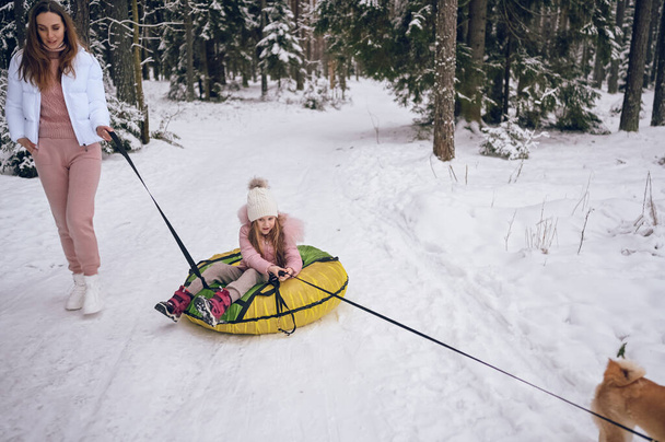 外で雪に覆われた白い冬の森の中で楽しい乗り物インフレータブルスノーチューブを持ってピンクの暖かい外を歩く幸せな母親と小さなかわいい女の子。家族スポーツ休暇活動 - 写真・画像