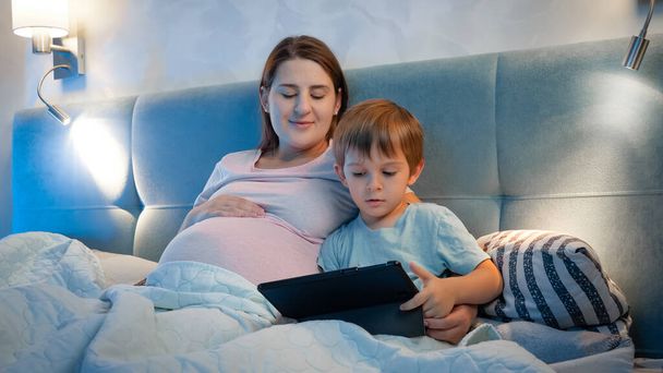 幸せな笑顔小さな男の子とともに妊娠中のお母さん見漫画上のタブレットコンピュータ上の夜にベッドで寝る前に - 写真・画像