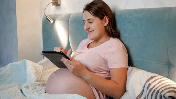 Koca göbekli, gülümseyen hamile bir kadının portresi. Yatağa yatmadan önce tablet bilgisayar kullanıyor.. - Fotoğraf, Görsel