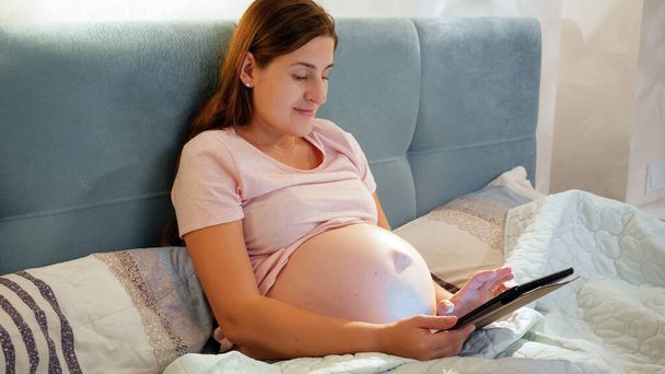 Lachende zwangere vrouw in pyjama met behulp van tablet computer en het typen van bericht terwijl ze in bed 's nachts - Foto, afbeelding