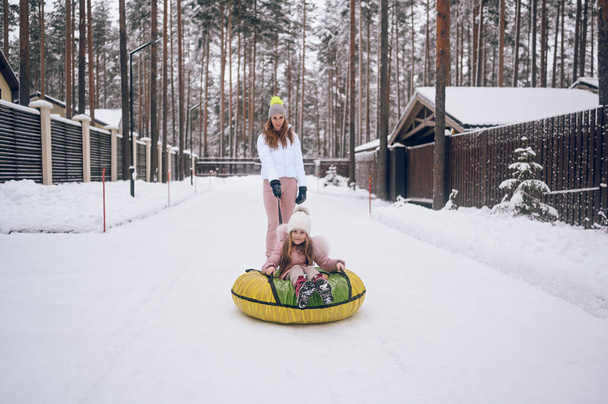 Happy mother and little cute girl in pink warm outwear walking having fun rides inflatable snow tube in snowy white cold winter forest outdoors. Family sport vacation activities - Photo, Image
