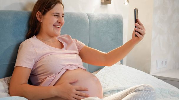 Femme enceinte souriante attendant bébé assis au lit et appelant un ami ou un médecin en vidéoconférence. Femme enceinte utilisant un smartphone - Photo, image