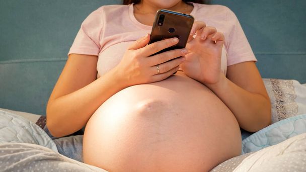 Closeup of pregnant woman with big belly in pajamas typing message on smartphone. Pregnant girl using smartphone in bed - Foto, imagen