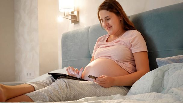Улыбающаяся беременная женщина делает онлайн покупки на планшетном компьютере перед сном ночью. - Фото, изображение