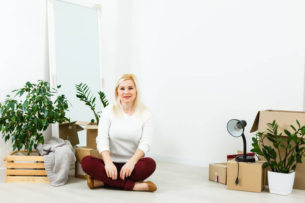 Happy young woman sitting on floor near moving boxes. Young woman moving to new home. Woman looking at camera and smiling - Photo, Image
