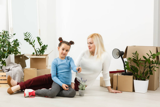 Cute mother and little daughter are on floor near boxes. They are preparing to move in another building. The parent and child are relaxing and smiling - Photo, Image
