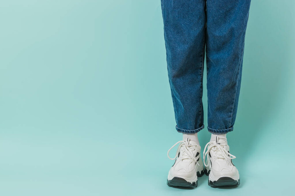 Legs of a girl in white socks, white sneakers and jeans on a blue background. Stylish youth clothing and shoes. Place for text. - Foto, afbeelding
