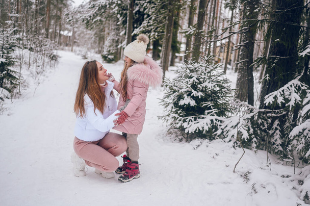 Happy mother and little cute girl in pink warm outwear walking having fun and hugging in snowy white cold winter coniferous forest with spruce woods outdoors. Family sport vacation activities - Photo, Image