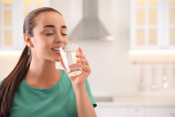 Young woman drinking pure water from glass in kitchen. Space for text - Fotó, kép