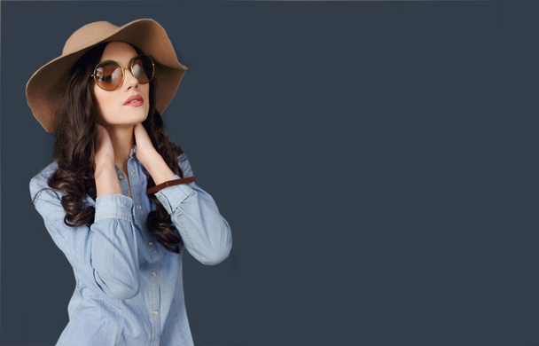 Αισθησιακό μελαχρινό κορίτσι με στρογγυλά γυαλιά ηλίου, σγουρά μαλλιά, φοράει τζιν πουκάμισο και μπεζ καπέλο, αγγίζει το λαιμό του. - Φωτογραφία, εικόνα