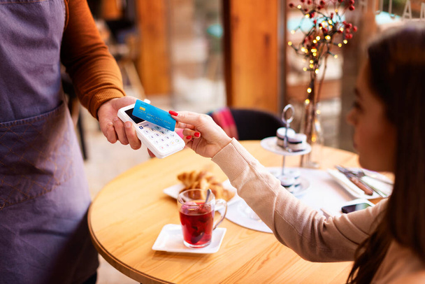 Ujęcie kobiety siedzącej przy biurku w kawiarni i płacącej kartą kredytową. Płatność zbliżeniowa.  - Zdjęcie, obraz