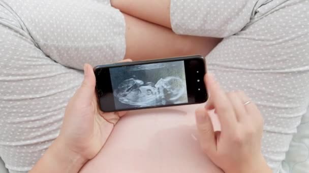 Κάτοψη της εγκύου που κάθεται στο κρεβάτι και κοιτάζει την εικόνα υπερήχων του αγέννητου μωρού της στο smartphone - Πλάνα, βίντεο