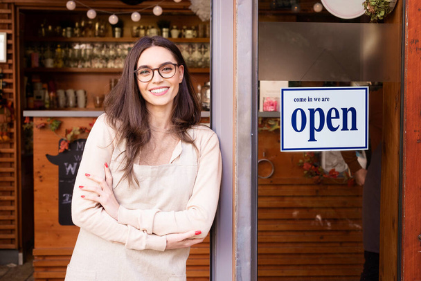 Shot of smiling young cafe show owner woman standing with arms crossed in the doorway. Open sign on the glass door. - Φωτογραφία, εικόνα
