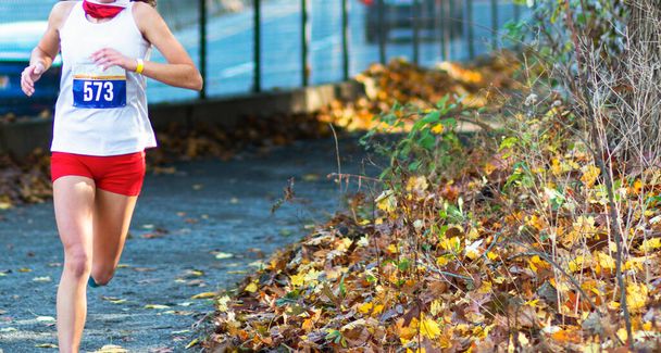 Licealistka biegnie w wyścigu biegowym po żwirowej ścieżce obok autostrady zablokowanej przez płot z jesiennymi kolorowymi liśćmi na poboczu ścieżki.. - Zdjęcie, obraz