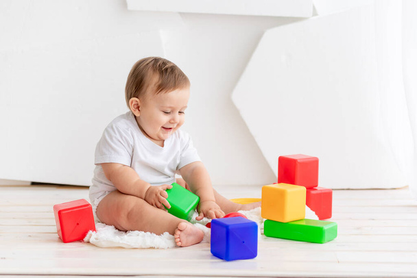 das Kind spielt, fröhliches kleines Baby sechs Monate alt in weißem T-Shirt und Windeln zu Hause auf einer Matte in einem hellen Raum mit bunten Würfeln - Foto, Bild