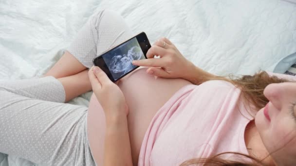 Νεαρή έγκυος γυναίκα με πιτζάμες, κοιτάζει υπέρηχο στο αγέννητο μωρό της. Έννοια της αναμονής του μωρού, της εγκυμοσύνης και της υγειονομικής περίθαλψης. - Πλάνα, βίντεο