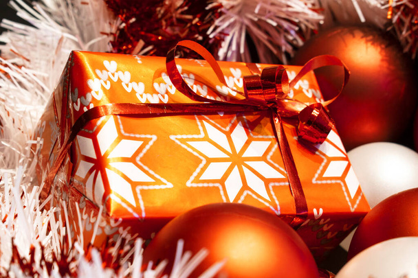 Рождественский подарок завернутый в красную декоративную бумагу среди украшений елки - Фото, изображение