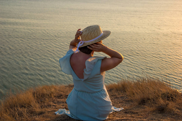 A girl in a dress and hat sits on a cliff by the sea - Photo, Image