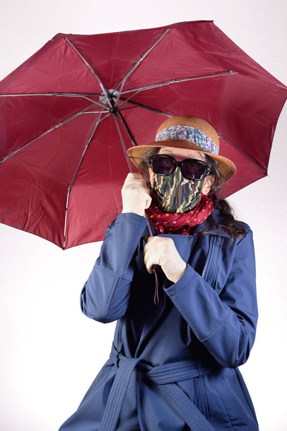 Woman protecting herself with umbrella and raincoat against the rain. The sunglasses and a straw hat protect her against the sun. The mask and the latex gloves against the covid-19 virus. Plain background. - Fotografie, Obrázek