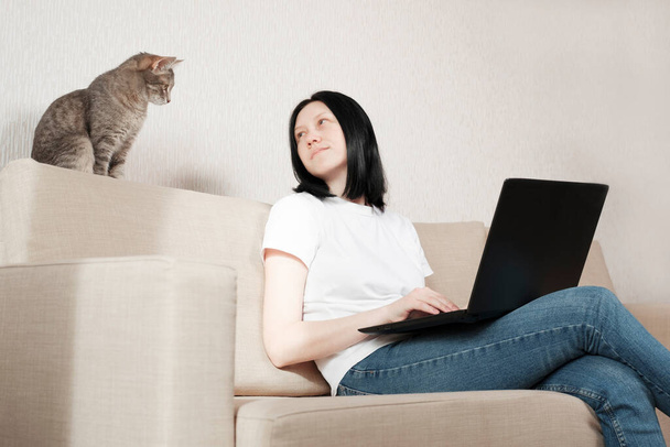 Een jonge vrouw werkt thuis terwijl ze op een bank zit. Het meisje gebruikt een laptop voor werk op afstand. Een grijze kat zit op de bank en volgt zorgvuldig het werk van de freelancer. Blijf thuis.. - Foto, afbeelding