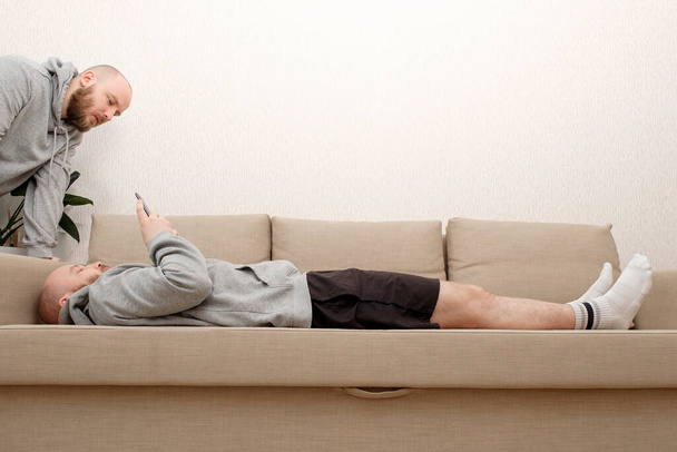 Collage de clonados masculinos se sienta en el sofá en varias poses y utiliza un teléfono inteligente. Compuesto de múltiples clones humanos que realizan diferentes actividades en casa durante el cierre debido a la pandemia de COVID - Foto, imagen