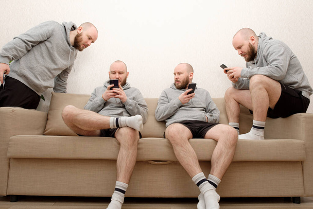 Collage de clonados masculinos se sienta en el sofá en varias poses y utiliza un teléfono inteligente. Compuesto de múltiples clones humanos que realizan diferentes actividades en casa durante el cierre debido a la pandemia de COVID - Foto, imagen