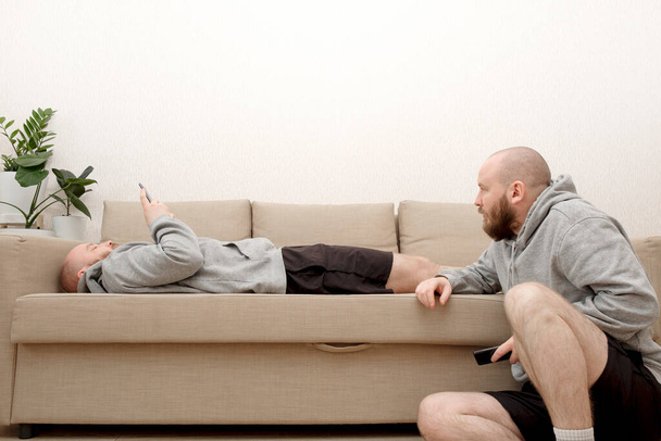 Collage de clonados masculinos se sienta en el sofá en varias poses y utiliza un teléfono inteligente. Compuesto de múltiples clones humanos que realizan diferentes actividades en casa durante el cierre debido a la pandemia de COVID - Foto, Imagen