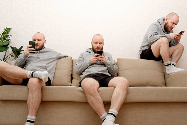 Collage of male cloned sits on the couch in various poses and uses a smartphone. Composite of multiple Human clones doing different activities at home during lockdown due to COVID pandemic - Fotoğraf, Görsel