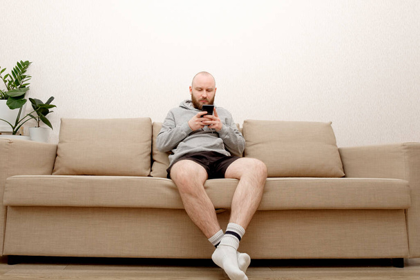 A young man with a beard in home clothes is sitting on a sofa and is using a smartphone. Smartphone - salvation from loneliness during self-isolation. The guy stayed home so as not to get a coronovirus. - Foto, Imagen
