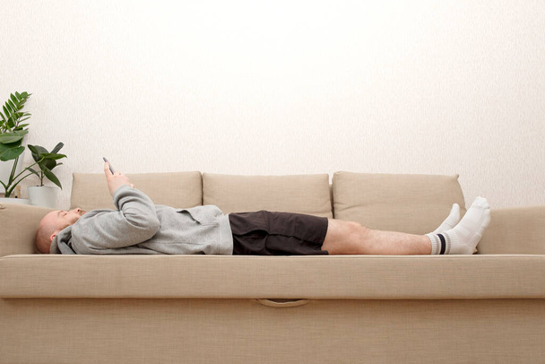 Młody człowiek z brodą w domowych ciuchach leży na kanapie i używa smartfona. Smartfon - zbawienie od samotności podczas samoizolacji.  - Zdjęcie, obraz