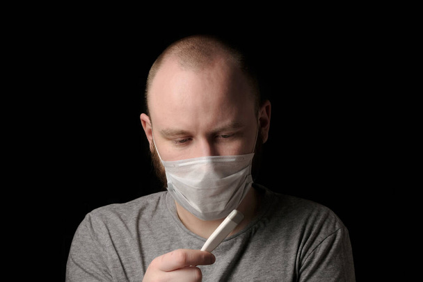 Egy beteg fiatalember orvosi maszkban feldúltnak tűnik, és egy lázmérőt tart a kezében. Megemelkedett testhőmérséklet. Szezonális fertőzés, megfázás, orrfolyás, koronavírus. Egészségügy és egészségügy - Fotó, kép