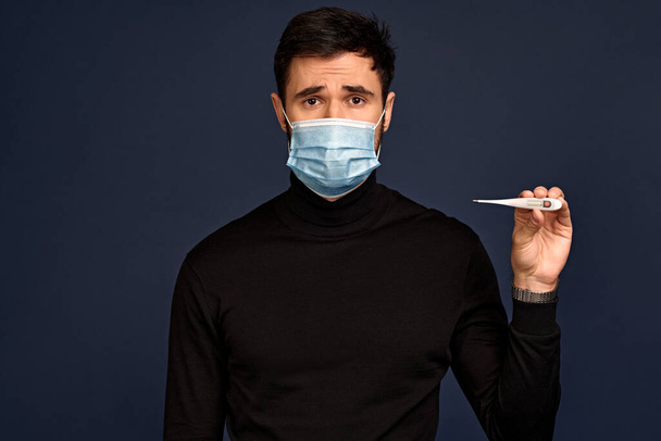 Mladý muž v černém svetru, se sterilní obličejovou maskou, která drží teploměr izolovaný na pozadí Pacific Blue ve studiu. Epidemie pandemie coronavirus 2019-ncov sars covid-19 chřipka virus koncept - Fotografie, Obrázek