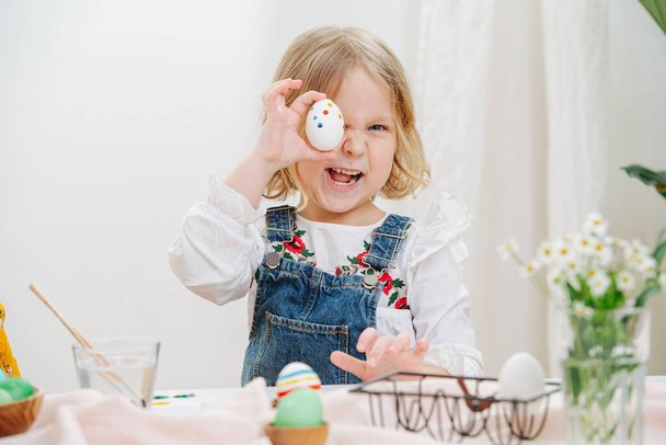 Маленькая девочка сидит за столом с пасхальными яйцами, держа один над глазом - Фото, изображение