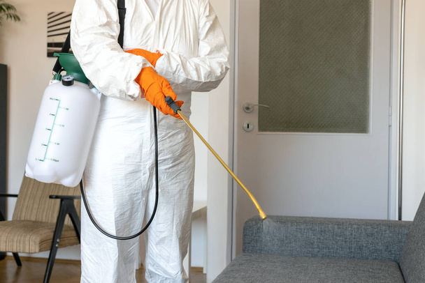 Persoon in beschermingspak met ontsmettingsspray fles desinfecterend huishouden en meubilair - Foto, afbeelding