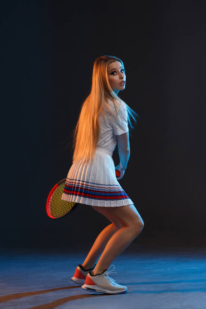 Όμορφη αθλητική κοπέλα αθλητής ξανθιά με μακριά μαλλιά παίζει τένις σε σκούρο φόντο με κίτρινο και μπλε πίσω φως. - Φωτογραφία, εικόνα