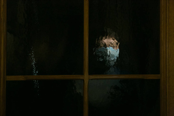 Καραντίνα στο σπίτι στο πρόσωπο του Coronavirus. Κλείδωμα. Το πρόσωπο μιας γυναίκας με μάσκα μέσα από σκούρο γυαλί. - Φωτογραφία, εικόνα