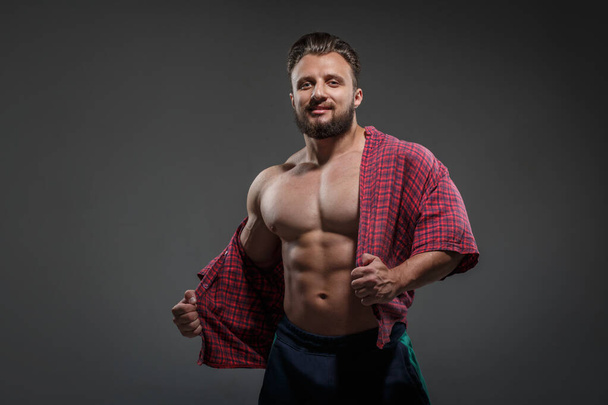 Muscular sexy white man takes off shirt isolated on dark background. - Foto, Imagen