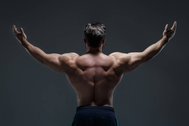 Portrait of a young athletic man with a naked torso showing muscles isolated on a dark gray background, view from the back. - 写真・画像