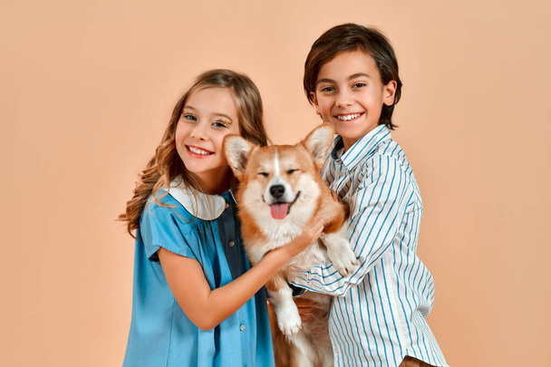 Το αγόρι στέκεται με την πλάτη του στην κάμερα, και ένα όμορφο κορίτσι με μπούκλες σε ένα φόρεμα κρατά ένα χαριτωμένο αστείο σκυλί στον ώμο της, απομονωμένο σε μια σκόνη ροδακινί φόντο. - Φωτογραφία, εικόνα