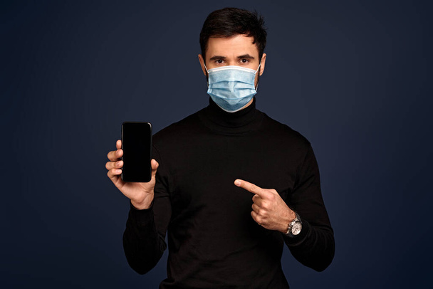 Young man in sterile face mask isolated on Pacific Blue background. Pandemic coronavirus 2019-ncov sars covid-19 flu virus concept. Pointing index finger on mobile phone with blank empty screen - Photo, Image