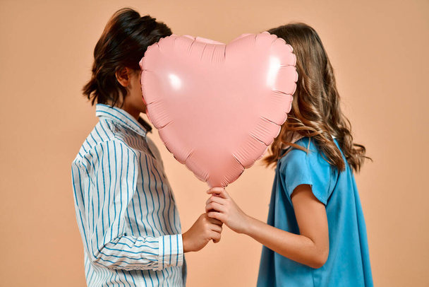 A cute lovely girl with curls in a blue dress and a nice boy in a shirt is holding a valentine heart balloon isolated on a powdery peach background. - Photo, image