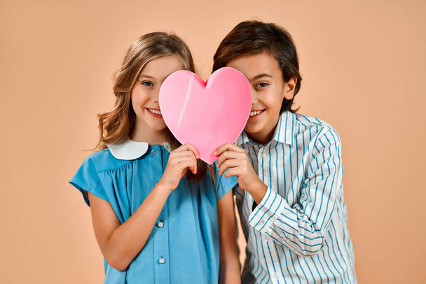 Cute lovely girl with curls in a blue dress and a nice boy in a shirt holding a valentine heart isolated on a powdery peach background. - Zdjęcie, obraz